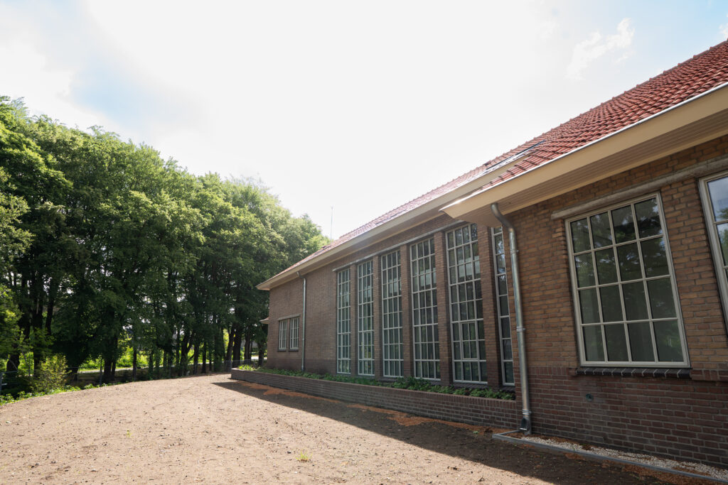 Lang bakstenen gebouw met grote ramen aan een grindpad langs de bomen