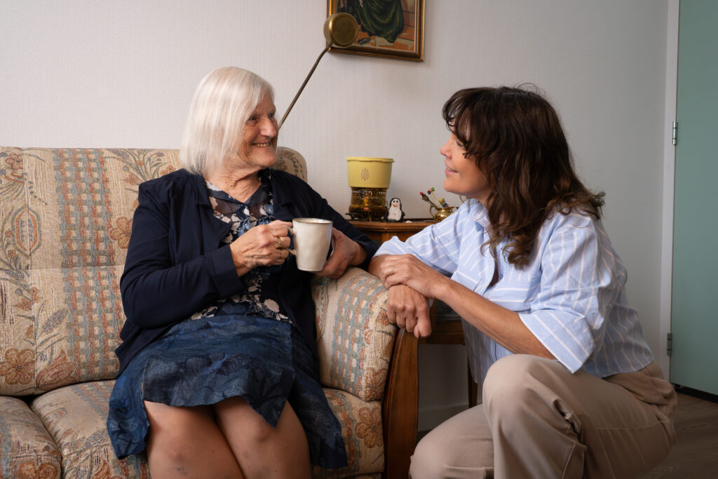 Oudere vrouw met een kop koffie op een gebloemde bank, in gesprek met een jonge vrouw naast haar